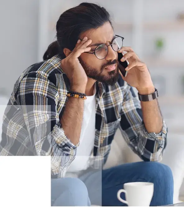 image of a long-haired man with a ponytail wearing flannel & eyeglasses touching one hand to his temple while talking on a smartphone - coffee cup on the table in front of him